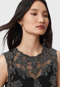 Jonge vrouw met lang, donker haar die kleine hoepelringen draagt en een zwarte mouwloze jurk met ingewikkelde zilveren parelmoer kanten bloemen.