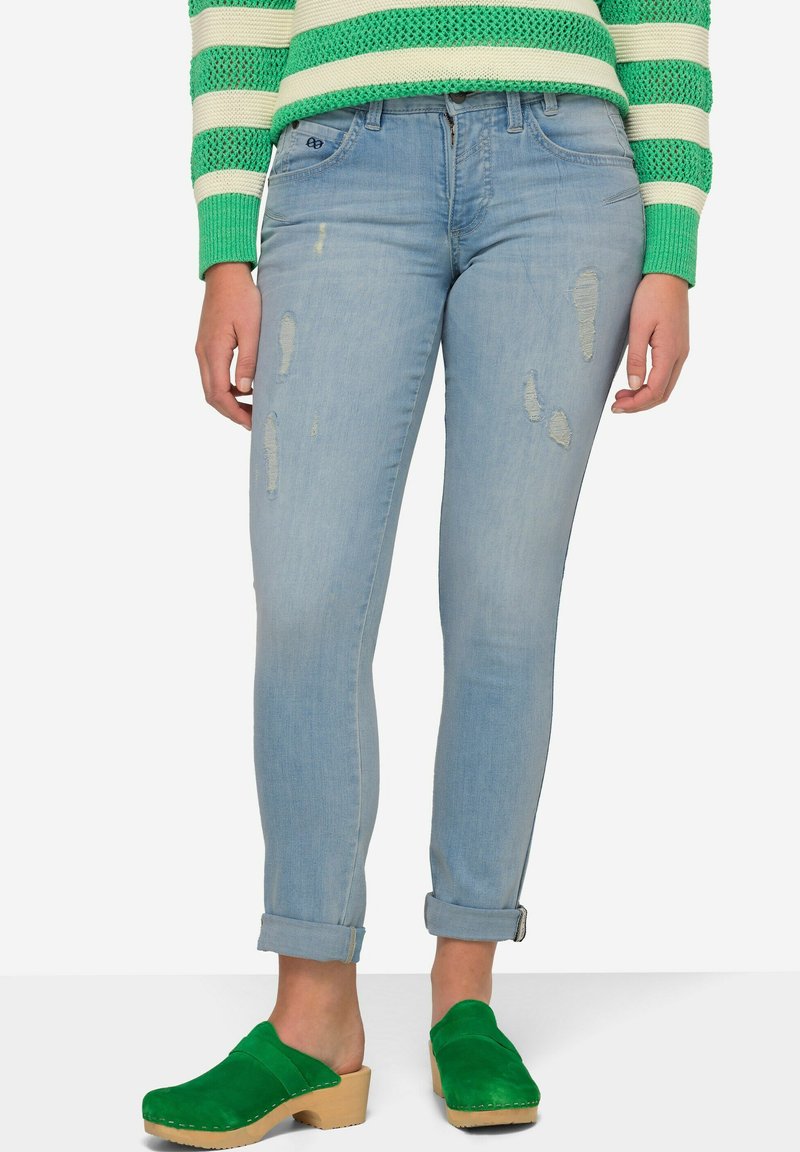 Jeans ajustados de mezclilla azul claro desgastados con puños enrollados, combinados con zuecos de ante verdes. Los jeans presentan patrones de desgaste visibles y detalles de costura.