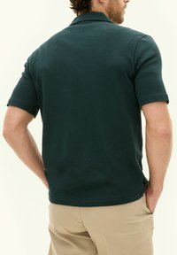 Tummanvihreä lyhythihainen polo-paita, jossa on perinteinen kaulus ja pehmeä kangas; yhdistettynä khaki-housuihin, näkyy takaa.
