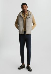 Helles graues gepolstertes Westchen mit Kapuze über einem sandfarbenen Poloshirt, dunkelblauen Hosen und braunen Slippern. Besitzt horizontale Nähte und eine Brusttasche.