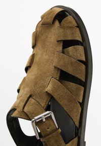 Sandalo in suede marrone con design a strisce intrecciate, rivestimento interno nero e fibbia metallica per una vestibilità regolabile. Sole piatto con bordi arrotondati.