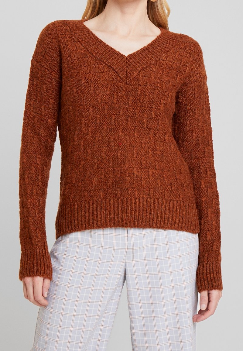 Femme portant un pull texturé col en V couleur rouille et un pantalon à carreaux gris clair sur un fond gris uni.