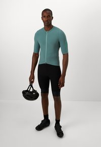 Koszulka kolarska z krótkim rękawem w kolorze teal z fakturowanym wzorem, z zamkiem z przodu, zestawiona z czarnymi szortami. Model trzyma czarny kask i ma na sobie buty kolarskie.