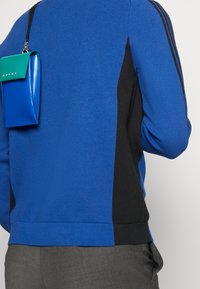 Maglione a maglia blu con pannelli laterali neri, dotato di una piccola borsa con zip verde e blu attaccata. Orlo e polsini a coste visibili.