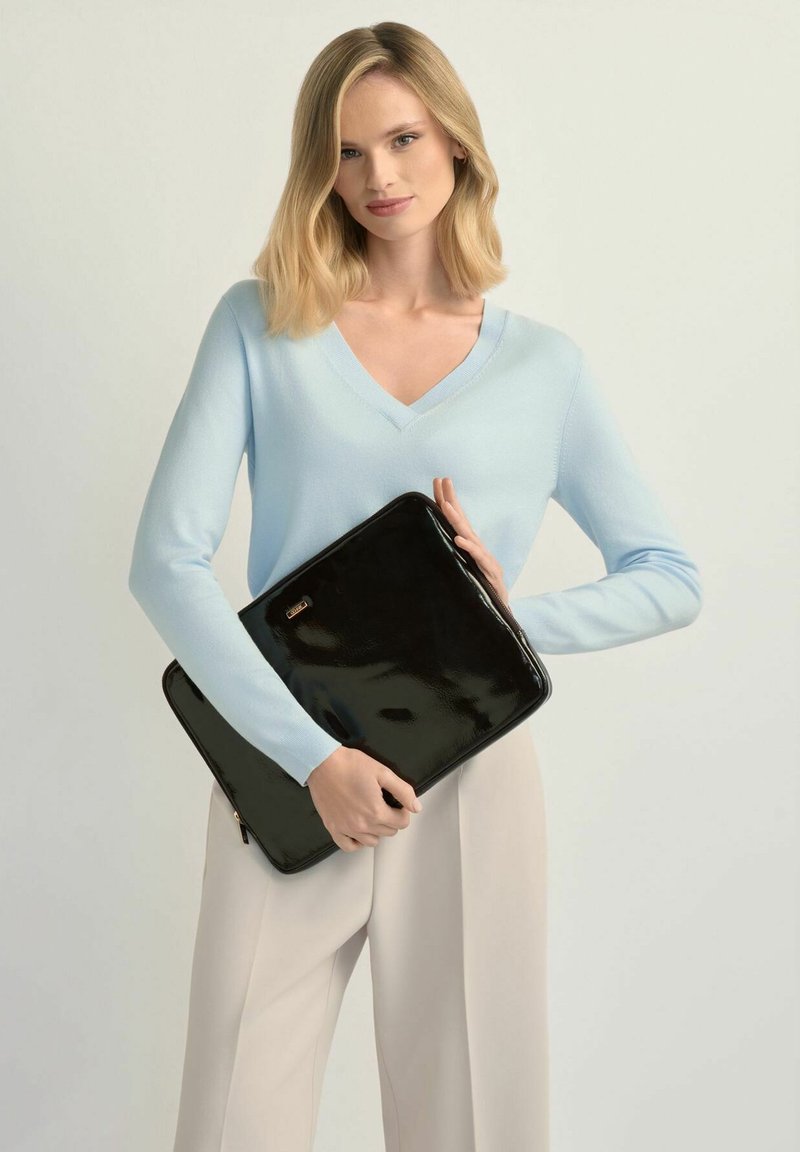 Blondynka w jasnoniebieskim swetrze z dekoltem w serek i beżowych spodniach trzymająca czarne błyszczące etui na laptopa na jednolitym tle.