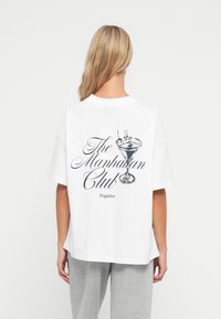 Balta kokvilnas T-kreklā ir melns drukāts dizains ar martini glāz�ē attēlu un tekstu "The Manhattan Club Pegador" uz muguras. Brīvs piegriezums.