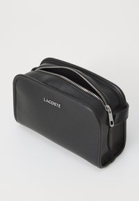Pochette da toeletta in pelle nera con texture a pietra, chiusura a doppia cerniera e manico laterale. Presenta un logo "LACOSTE" in argento.