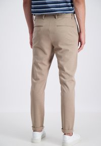 Muž v béžových slim-fit kalhotách s ohrnutými lemy, bílých teniskách a modrém pruhovaném overshirtu, stojící proti jednoduchému světlému pozadí.