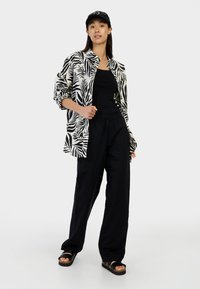 Witte shirt met zwart bladenpatroon, zwarte tanktop, losse zwarte broek en zwarte sandalen. Zwarte pet gedragen op lang, steil haar.