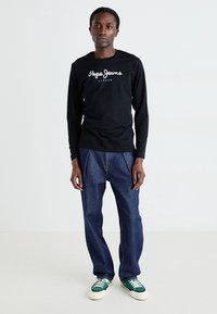 Černé tričko s dlouhým rukávem a bílým logem "Pepe Jeans London", spárované s vysokými modrými džínami a zelenými teniskami.