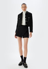 Veste noire courte avec des boutons argentés, assortie à une jupe enroulée noire ajustée. Le mannequin porte un col roulé blanc et des chaussures à plateforme noires avec des accents.