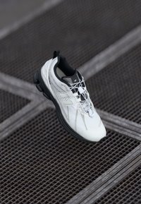 Scarpa sportiva bianca con tomaia testurizzata, suola nera e dettagli riflettenti. Presenta un collo aderente e lacci elastici per una vestibilità sicura.