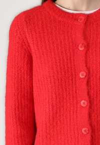 Cardigan en tricot rouge avec une texture côtelée, comportant six boutons roses et un col rond. Le tissu semble doux et chaud.