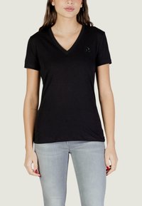 Młoda kobieta ubrana w czarny T-shirt z krótkim rękawem i dekoltem w serek, z małym wyszywanym literą "A" na lewej piersi, w połączeniu z jasnoszarymi dżinsami.