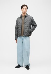 Homme portant une veste matelassée grise, un pull marron, un jean large bleu clair et des chaussures noires, debout avec une main dans la poche.