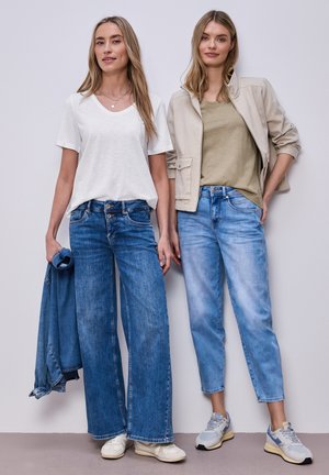 Dwie kobiety stoją obok siebie w codziennych ubraniach: jedna w białym t-shircie i szerokich dżinsach trzyma kurtkę dżinsową, druga w beżowej kurtce i jasnych dżinsach.