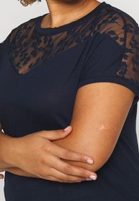 Mörkblå t-shirt med en genomskinlig spetspanel med blommönster på axlarna och övre bröstet. Kort ärm och rundad halslinje.