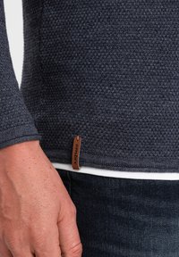 Marineblauer strukturierten Pullover mit langen Ärmeln, mit einem Lederetikett am Saum. Getragen über einem weißen Hemd und dunklen Jeans.