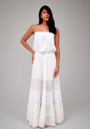 Vrouw met lang donker haar die een strapless witte maxi-jurk met kanten details draagt, staand tegen een effen grijze achtergrond.