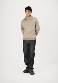 Sudadera de forro polar beige con cremallera y cuello alto, combinada con jeans oscuros de corte holgado y zapatillas negras de suela gruesa.