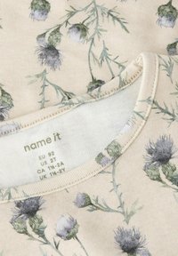 Tela con estampado floral que presenta diseños de cardos en púrpura y verde sobre un fondo beige; escote redondo con bordes cosidos.