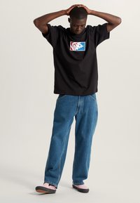 Camiseta negra de manga corta con el logo de Vans en rojo y azul, combinada con jeans de mezclilla azul y zapatillas de slip-on en negro y rosa.