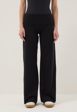 Donna che indossa pantaloni neri a gamba larga e scarpe slip-on bianche con suole color cuoio, in piedi davanti a uno sfondo chiaro e semplice.