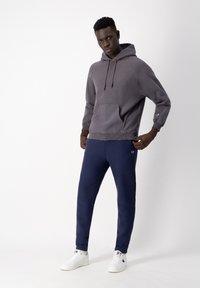 Gray hoodie with a front pocket and drawstrings, paired with navy sweatpants. White sneakers and subtle logo accents are visible.