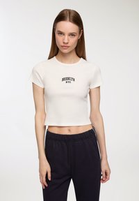 Biała krótka koszulka z ribbowanej tkaniny z napisem "BROOKLYN NYC" na czarnym tle. W zestawie z ciemnymi spodniami dresowymi, prezentującymi luźny krój.