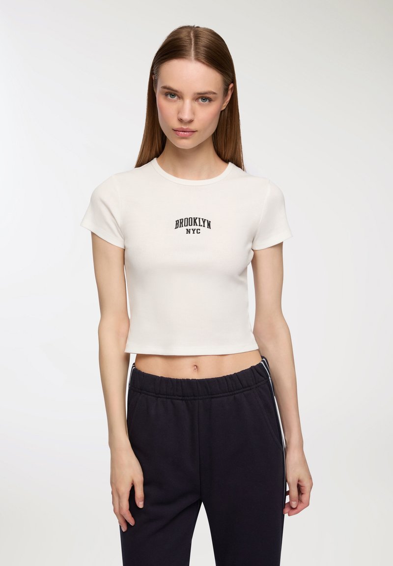 Biała krótka koszulka z ribbowanej tkaniny z napisem "BROOKLYN NYC" na czarnym tle. W zestawie z ciemnymi spodniami dresowymi, prezentującymi luźny krój.