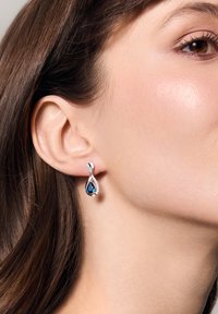 Niebieskie kolczyki w kształcie kropli łzy z srebrnymi akcentami, posiadające skomplikowane wzory oraz małe, przezroczyste kamienie wzdłuż obręczy, które kontrastują z odcieniem skóry.