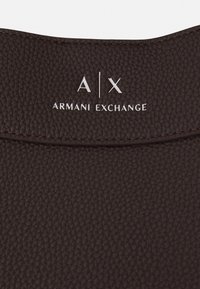 Pelle marrone testurizzata con logo "A|X ARMANI EXCHANGE" in bianco, posizionato al centro su uno sfondo scuro.