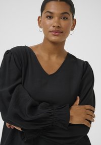 Schwarze V-Ausschnitt-Bluse mit Puffärmeln und gerafften Bündchen, aus leichtem Textil mit glatter Struktur. Ausführliches Design rund um den Halsausschnitt.
