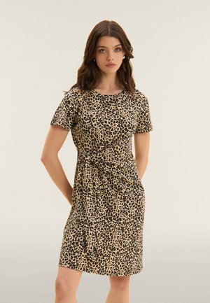 Donna con capelli castani ondulati che indossa un vestito corto con maniche corte e stampa leopardata, lungo fino al ginocchio, in piedi con le braccia dietro la schiena, davanti a uno sfondo semplice.