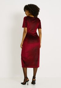 Robe midi en velours rouge avec manches courtes, coupe ajustée et texture subtile à côtes verticales. Associée à des chaussures noires à talons hauts.
