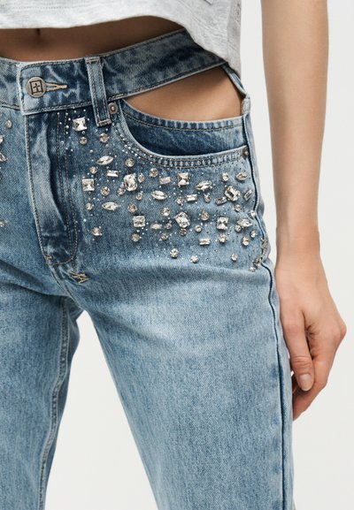 Τζιν τζιν με διακοσμητικές κρυσταλλικές λεπτομέρειες στις μπροστινές τσέπες και μια ορατή λεπτομέρεια κοπής κοντά στη μέση, φορεμένα από ένα άτομο.