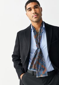 Braun-blau gemusterter Seidenschal locker um den Hals eines Mannes gebunden, getragen über einem hellblauen Hemd und einem schwarzen Mantel, mit dezentem Glanz und weicher Textur.