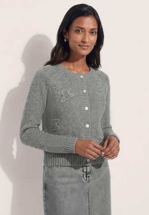 Frau mit dunklen Haaren, die eine graue, geknöpfte Strickjacke und einen passenden grauen Jeansrock trägt, steht vor einem einfarbig hellen Hintergrund.