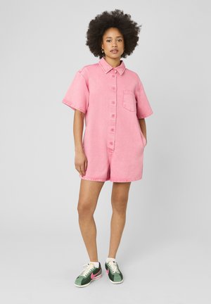 Femme avec coiffure afro portant une combinaison courte rose à boutons et des baskets vertes avec des touches roses, debout devant un fond uni.