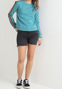Felpa turchese con strisce arancioni sulle maniche, abbinata a pantaloni corti di jeans scuri e sneakers nere. Tessuto morbido, vestibilità casual, senza loghi visibili.