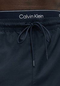 Melnas peldbikses ar elastīgu vidukļa jostu un sasietu aukliņu, ar Calvin Klein logotipu uz iekšējās vidukļa jostas.