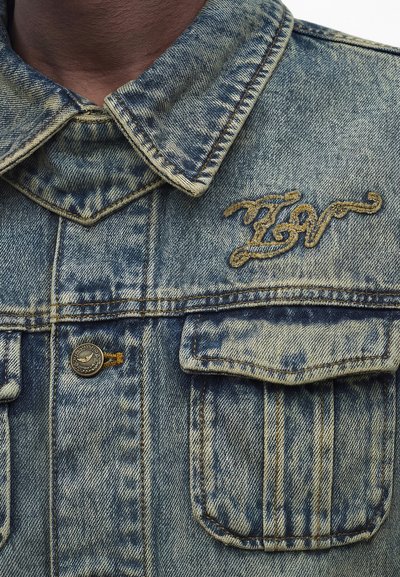 Τζιν μπουφάν με τσέπες με καπάκι και κουμπιά και κίτρινη κεντημένη επιγραφή στο στήθος κοντά στον γιακά.