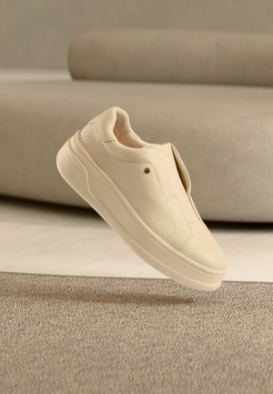 Μπεζ sneaker χωρίς κορδόνια με υφασμένο δερμάτινο επάνω μέρος και παχιά σόλα να αιωρείται πάνω από μπεζ χαλί, με έναν θολωμένο μπεζ καναπέ στο φόντο.