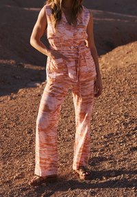 Combinaison à motifs roses et orange avec un col, une patte de boutonnage à l'avant, une ceinture à nouer et des jambes larges. Portée avec des sandales marron sur une surface texturée.