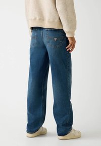 Jeans in denim blu con una vestibilità rilassata, caratterizzati da due tasche posteriori e dettagli di cucitura sottili. Indossati con un maglione beige chiaro e scarpe beige.