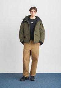 Pufasta jakna maslinasto zelene boje preko crne majice, u kombinaciji s bež hlačama i crnim tenisicama, s opuštenim krojem i jednostavnim dizajnom.