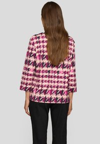 Frau mit langem braunem Haar, die eine Jacke im Hahnentrittmuster in Pink, Lila und Schwarz sowie eine schwarze Hose trägt, von hinten gesehen.