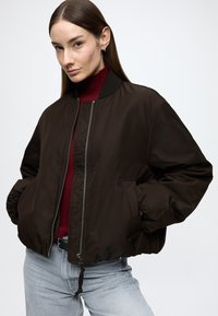 Mørk brun bomberjakke med glidelås foran, ribbet krage og elastiske mansjetter. Har sidelommer og et teksturert fôr. Kombinert med en rød skjorte.