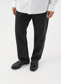 Svarta jeans med blanka svarta loafers. Skjortan är vit med tunna vertikala ränder, har långa ärmar och en knäppt krage.