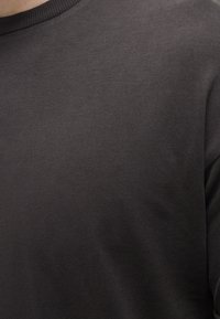 Gros plan d'une personne portant un pull uni gris foncé à col rond, montrant la partie supérieure de la poitrine et les épaules.